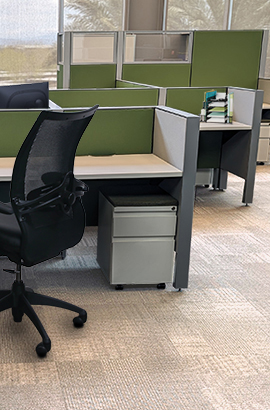 Modern open-plan office with cubicles featuring olive green fabric partitions, light grey desk surfaces, black ergonomic mesh office chairs, and large windows providing natural light.
