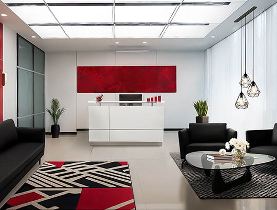 Modern office reception area with white reception desk, black modern sofas and armchairs, red accent wall, geometric patterned rugs, and glass coffee table with decorative elements.