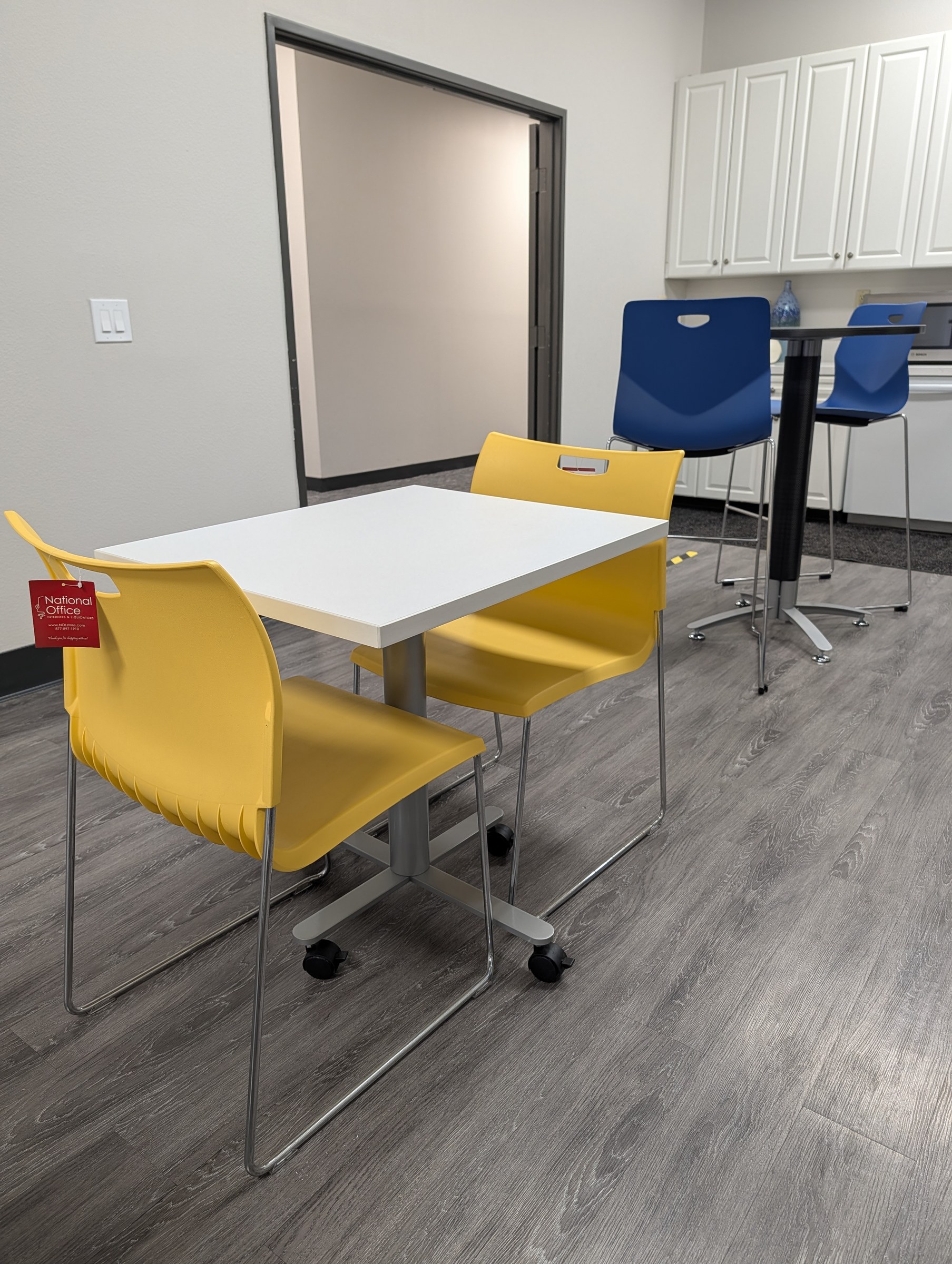 Breakroom setup with a white cafe table, yellow stack chairs and a bar height table with blue stools